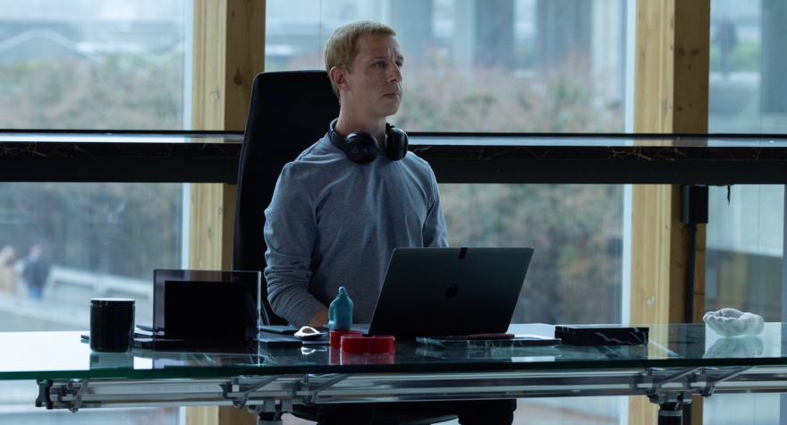 A man wearing headphones around his neck sits at an expansive desk in an office with huge windows