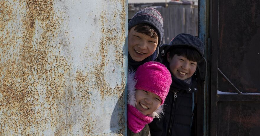 Three children wearing hats and scarves peek round a wall, smiling.