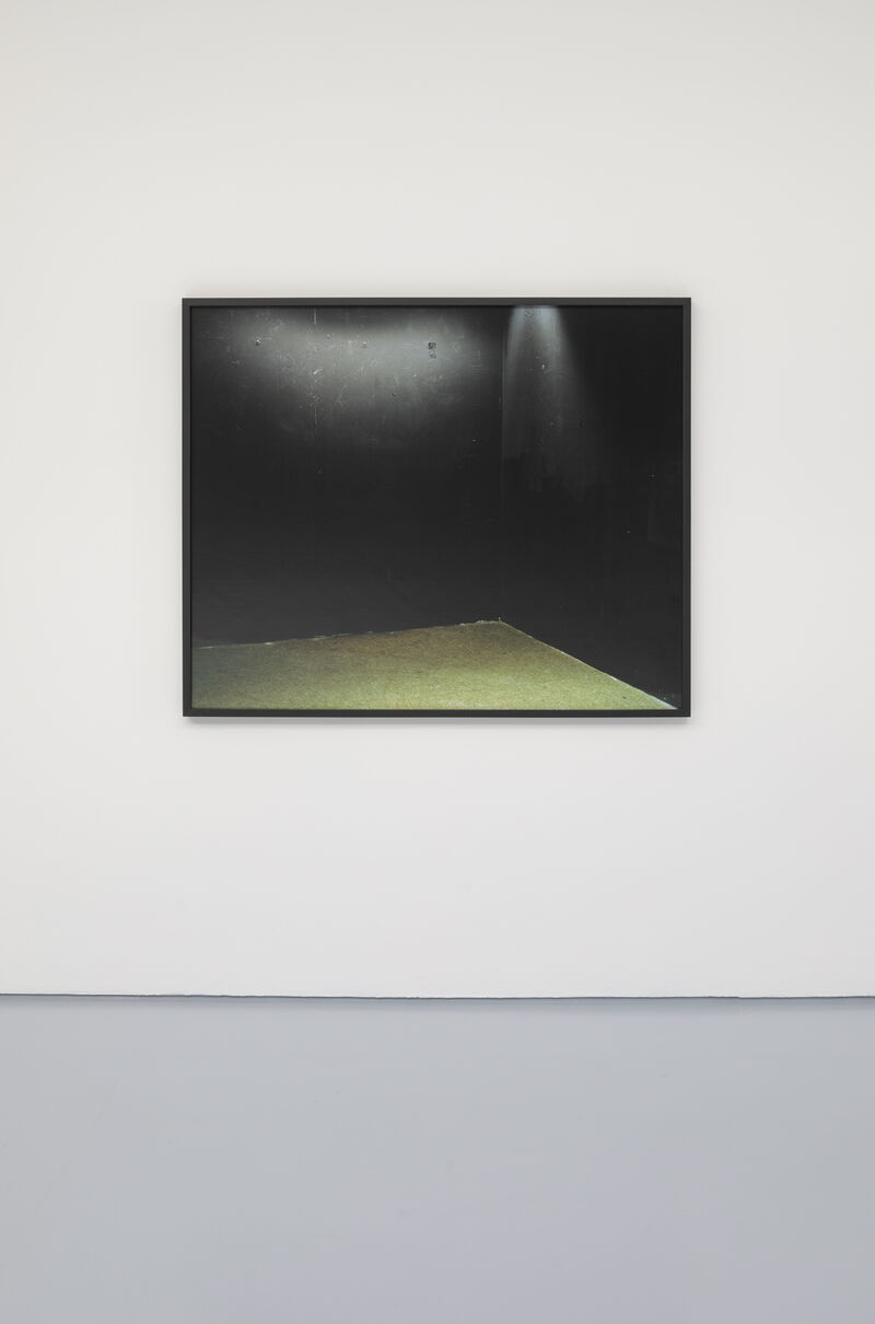 A photograph in a slim black frame is hung on a white wall. In the photograph we can see the corner of a black room with a spotlight in the corner, as though an empty stage. 