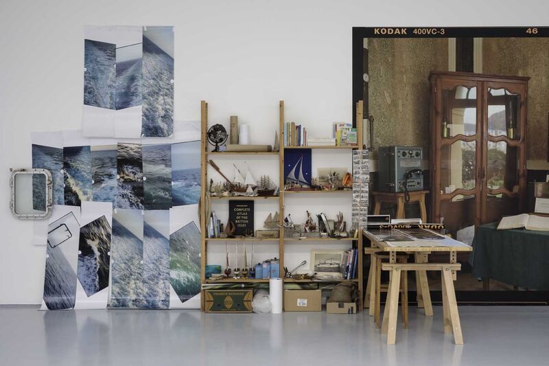 A gallery wall, against which are several photographs of the sea, shelves containing ephemera including model ships and shells.