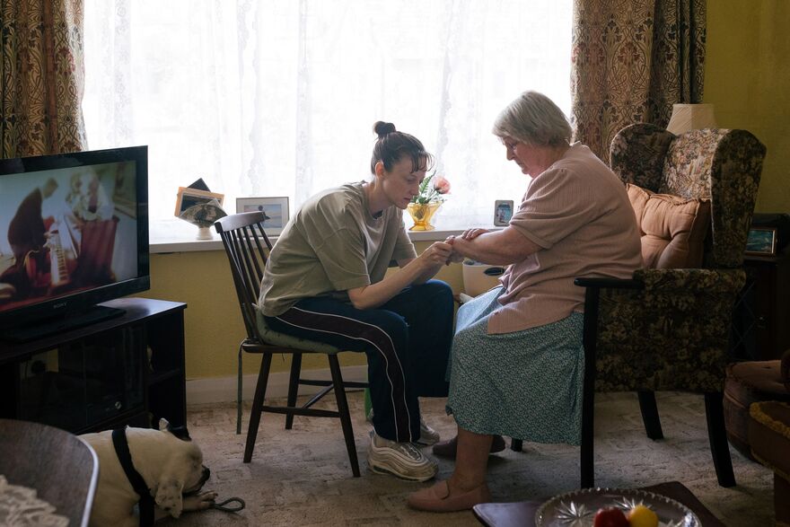 An elderly woman and a younger woman in a tracksuit sit in a living room. The younger woman looks at the older woman's wrist
