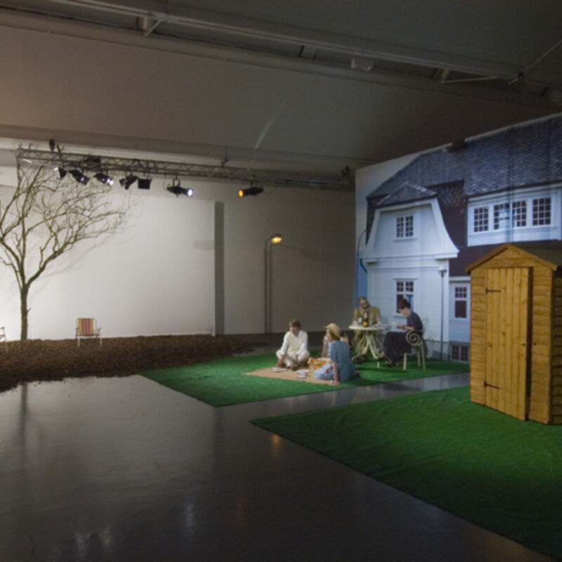 From Killing Time at DCA Galleries. Four people have a picnic on fake gras in the galleries in front of a large fabric sheet with a photograph of a house on it. There is a real wooden shed next to the people, along with a tree with no leaves, and two deckchairs.