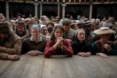 A crowd of medieval people wearing medieval clothing stand in a theatre. A woman wearing a red dress in the centre of the image puts her elbows on the stage and clasps her hands.