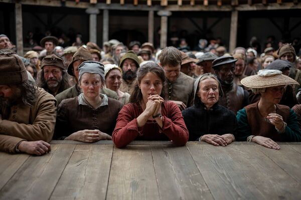 A crowd of medieval people wearing medieval clothing stand in a theatre. A woman wearing a red dress in the centre of the image puts her elbows on the stage and clasps her hands.