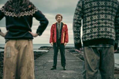 A man in a red jacket is stood facing us and the back of a woman and of a man stand infant of us framing the man in the red jacket
