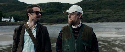 Two middle-aged men stand outside in a scenic location filled with trees. They smile at each other knowingly.