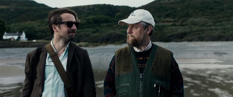 Two middle-aged men stand outside in a scenic location filled with trees. They smile at each other knowingly.