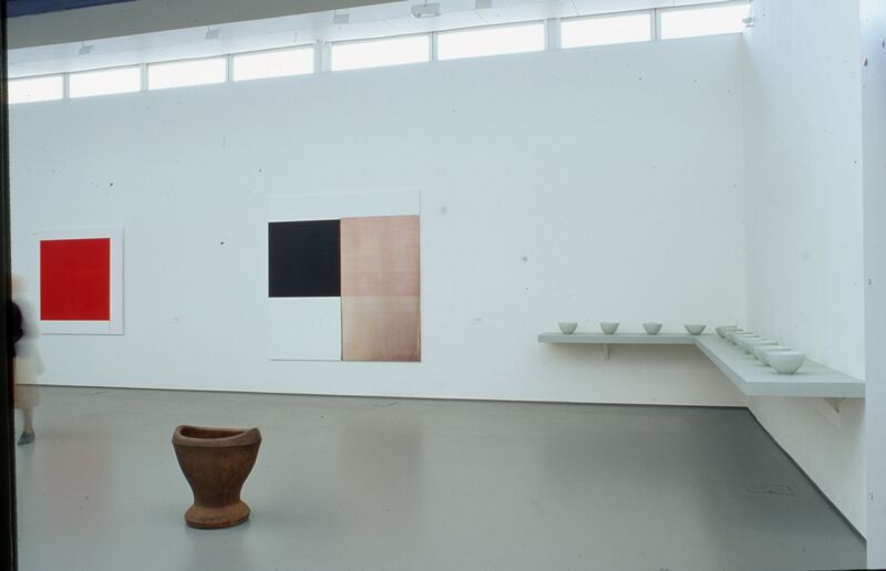 Installation view of the Prime exhibition at DCA - a shelf holds identical white ceramic bowls, and a large ceramic vase. There are also single colour geometric prints on the white walls.