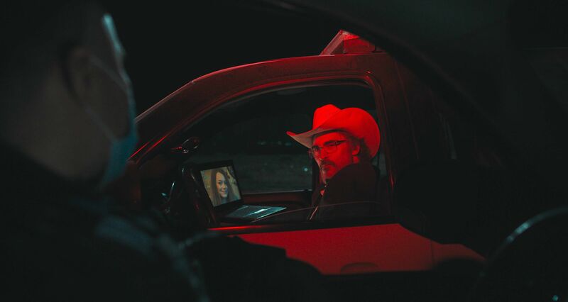 A man wearing a cowboy hat sits in a car, lit with red light. A woman's face is on his laptop screen which is sitting on the dashboard.