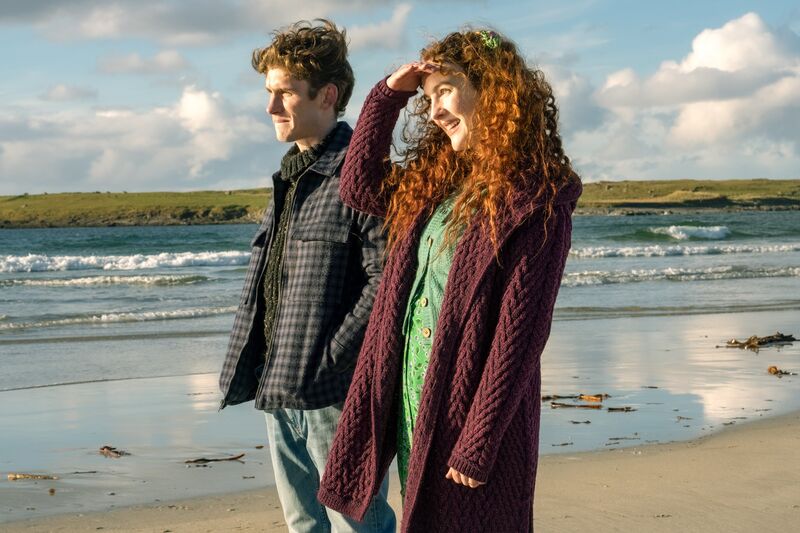 A young man and woman in knitwear stand on a beach smiling.