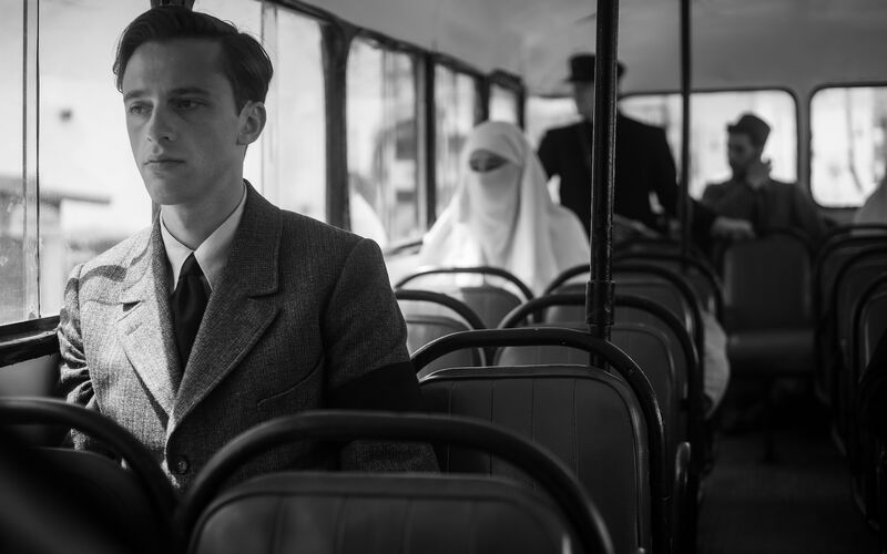 A black and white still of a scene on a bus, a white man sits near the front and a lady sits a few rows behind wearing a Burka 