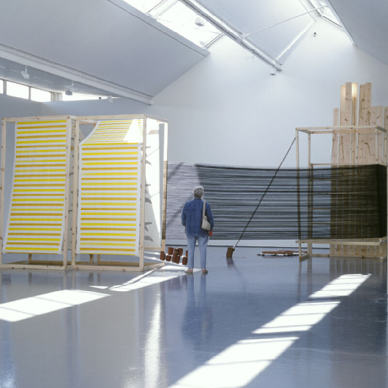 From Claire Barclay's exhibition. A person stands in DCA Galleries looking at Claire Barclay's sculptures. One is a large plywood structure with pink and yellow stripes painted on it. There is also semi-transparent black material, and wooden beams.