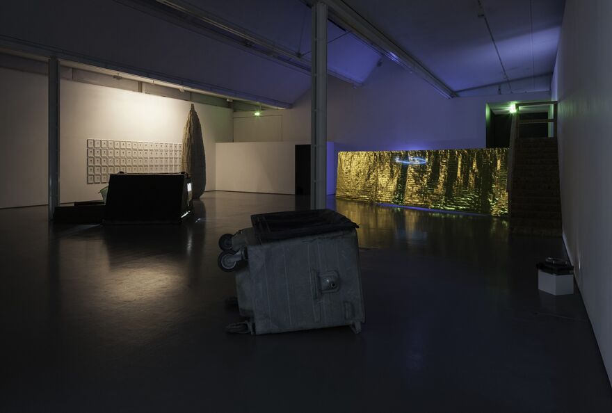 A gallery view from Navid Nuur's exhibition. A large bin is toppled over on its side. In the background, there is a large metallic sheet.