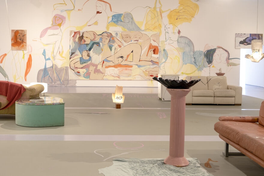 a gallery wall with figures painted on it in. In front of the gallery are sofas and a pedestal 