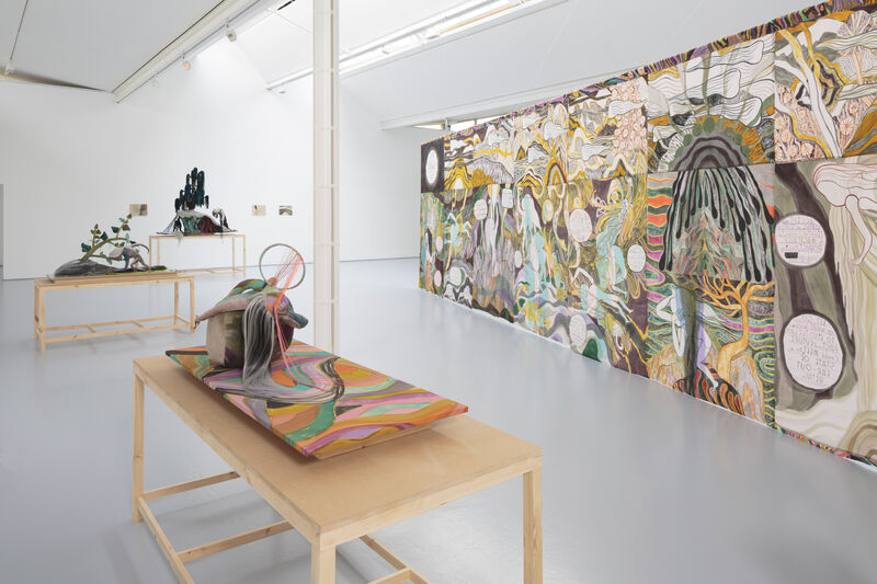 To the left hand side are three plinths with a textile artworks. A hung textile work with watercolour painting and graphic text hangs to the right. The colours are muted and soft, the gallery is naturally lit and bright. 