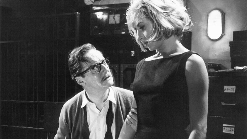 A black-and-white film still shows a nervous man holding a woman's arm