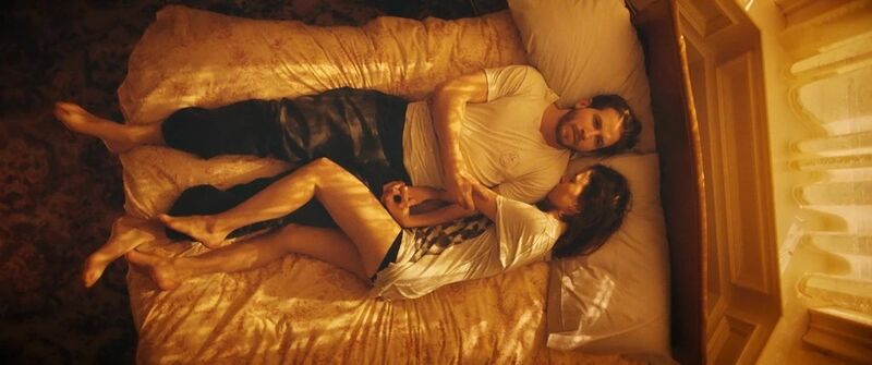 A man and woman lie on a bed together. The woman lies on her side cuddling into the man, they both have neutral expressions.