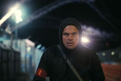A man stands in a dark street wearing a dark hoodie with the hood up.