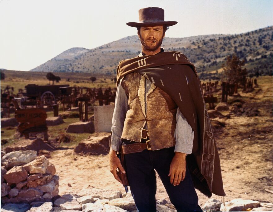A man stands in a poncho and hat, smoking a cigar in the Old West