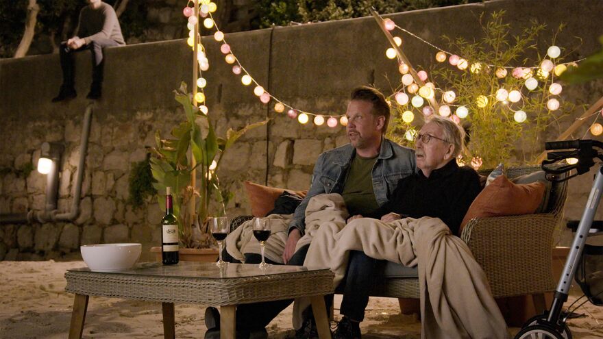 A man and his elderly father sit outside with glasses of wine.
