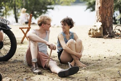 A man and woman in summer clothing sit by a lakeside smiling at each other.