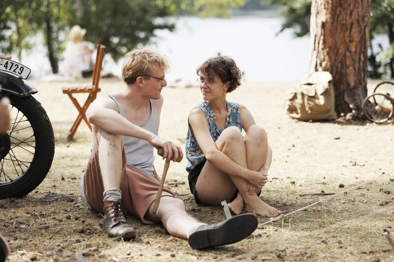 A man and woman in summer clothing sit by a lakeside smiling at each other.