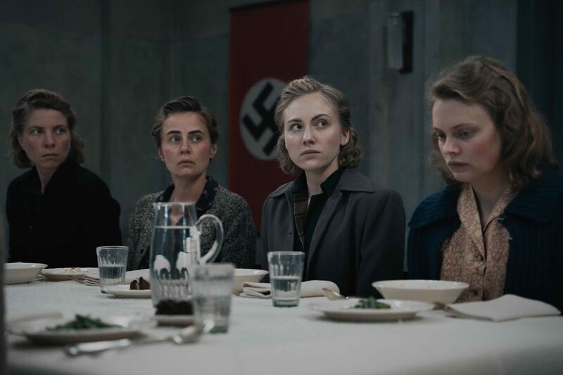 A group of solemn looking woman sit at a dinner table with a Nazi flag behind them