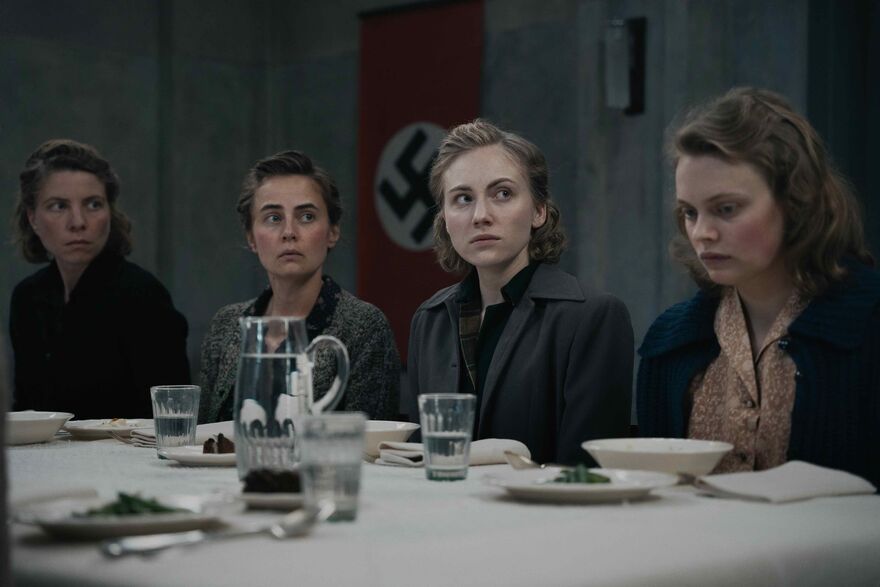 A group of solemn looking woman sit at a dinner table with a Nazi flag behind them