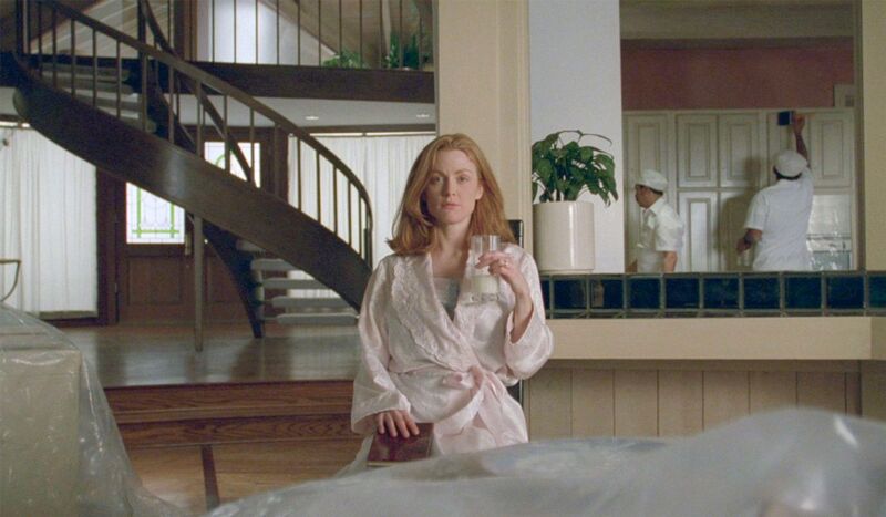 A woman in a pink dressing gown sits in the centre of the a very tidy house scene. A spiral staircase and cleaners in white visible in the background