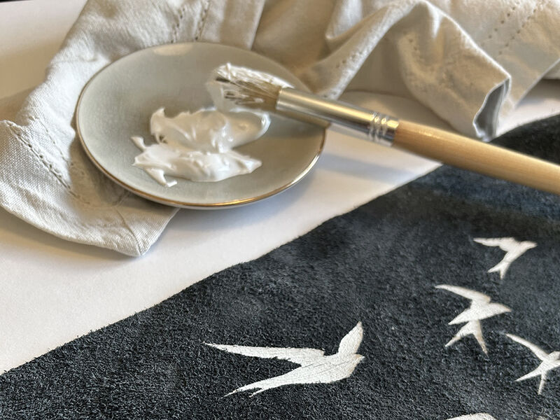 Paint brush resting on a plate with white ink. Beside it is a piece of black suede leather with a bird design on it in white ink