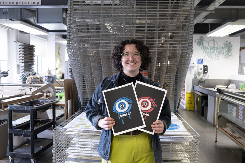 Illustrator Katrion Gillon holding two Dundead 2025 screenprints, one red, one blue