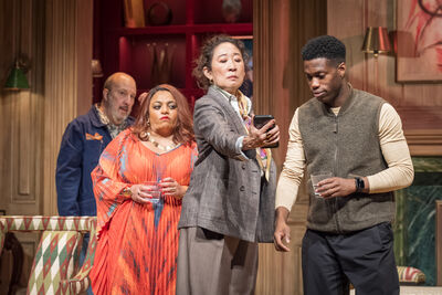A stage setting a lady looks at her phone with a man while two people in the background try to peer round to see it