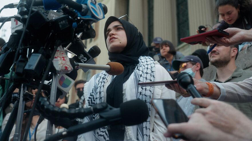 A young woman with a Palestinian scarf round her shoulders speaks to a group of reporters.