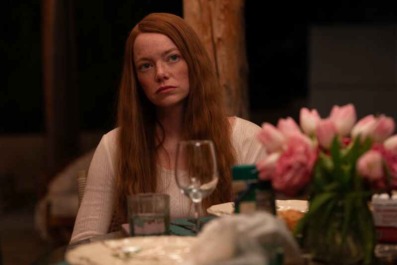 A woman with long red hair wearing a white shirt sits a table with pink tulips on it.