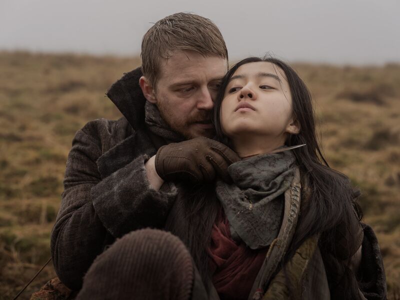 In a marsh, a man holds a knife to a young woman's throat.