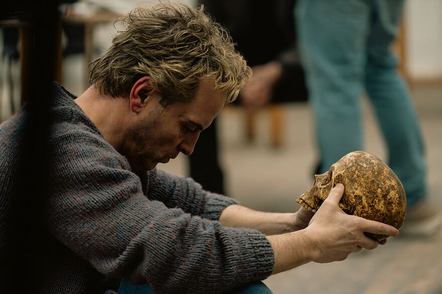 A man crouches with his head bowed, holding a skull in his hands