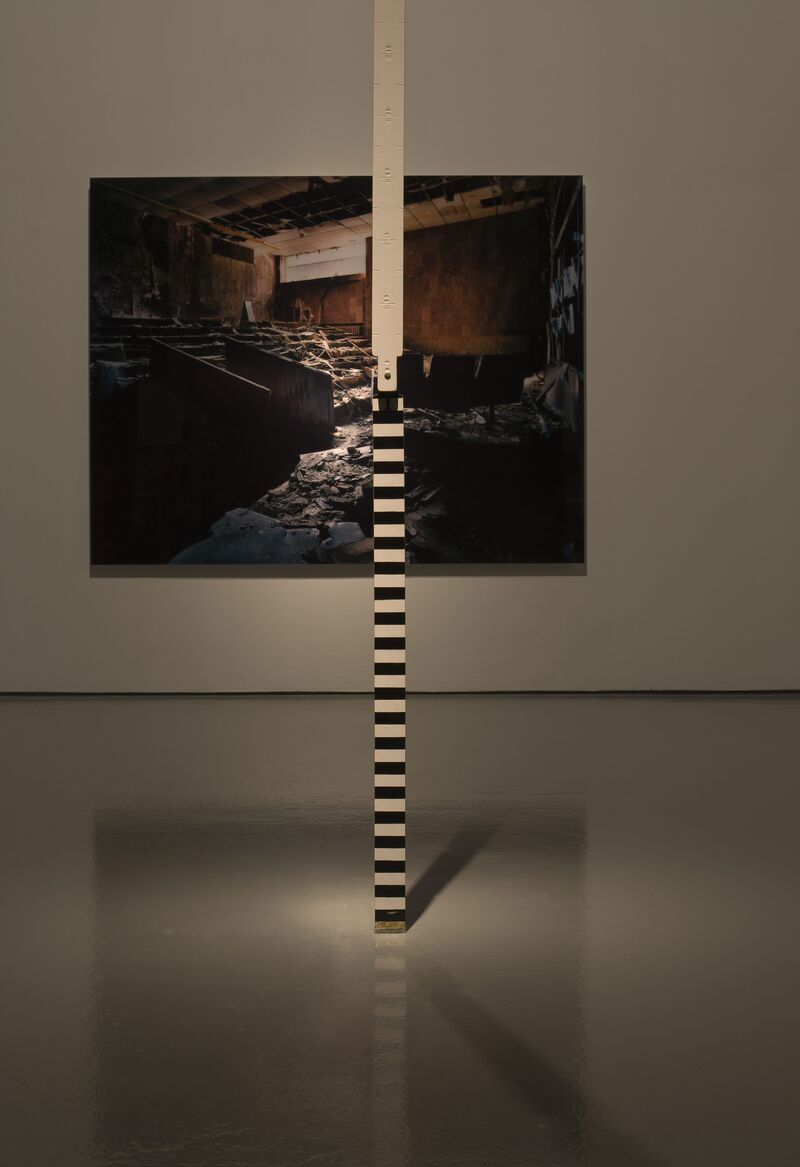 From Jane & Louise Wilson's exhibition. A large pole is erected in the gallery. It is painted with black and white stripes. A photograph of an abandoned and decaying room is hung on the gallery wall behind.