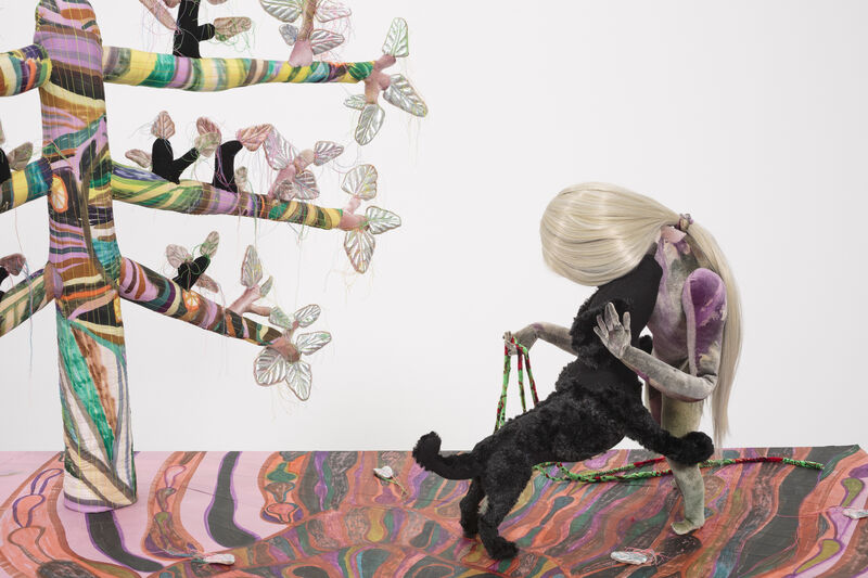 A close up of a textile sculpture, with a figure bending down to a black dog on the right. On the left is a tree. The fabric pieces are mounted on a painted board which is mainly pink and red. 