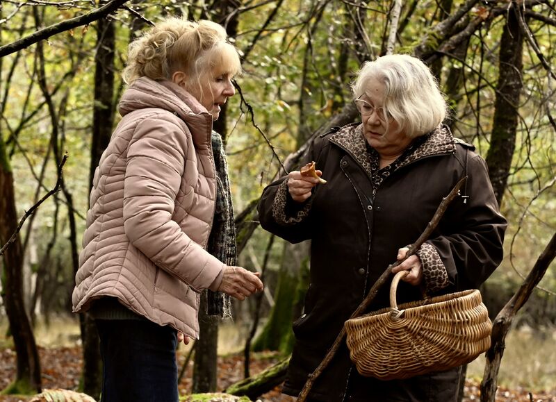Two older woman stand in a forest, holding a basket.