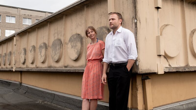 A man in a white shirt leans against a brutalist building smoking, while a woman in an orange dress looks at him smiling.