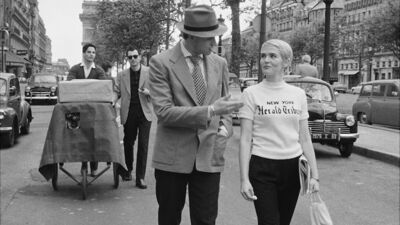 A black-and-white film still shows a man wearing a suit and a hat and a woman with a short haircut walking down a Paris street in the 1960s.