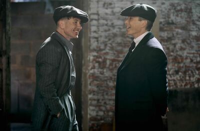 Two men in forties-style hats and suits look at each other laughing