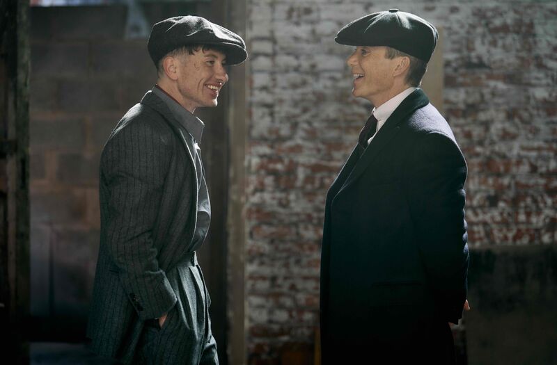 Two men in forties-style hats and suits look at each other laughing