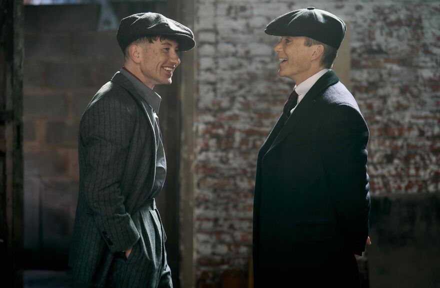 Two men in forties-style hats and suits look at each other laughing