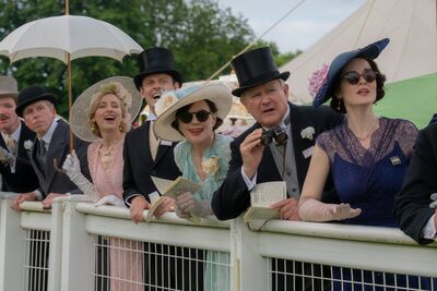 A group of people in formal, colourful dresses and tophats watch a race.