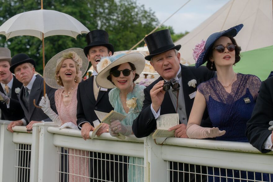 A group of people in formal, colourful dresses and tophats watch a race.