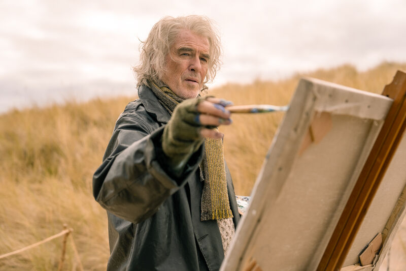 A man with long grey hair stands on a beach painting outside.