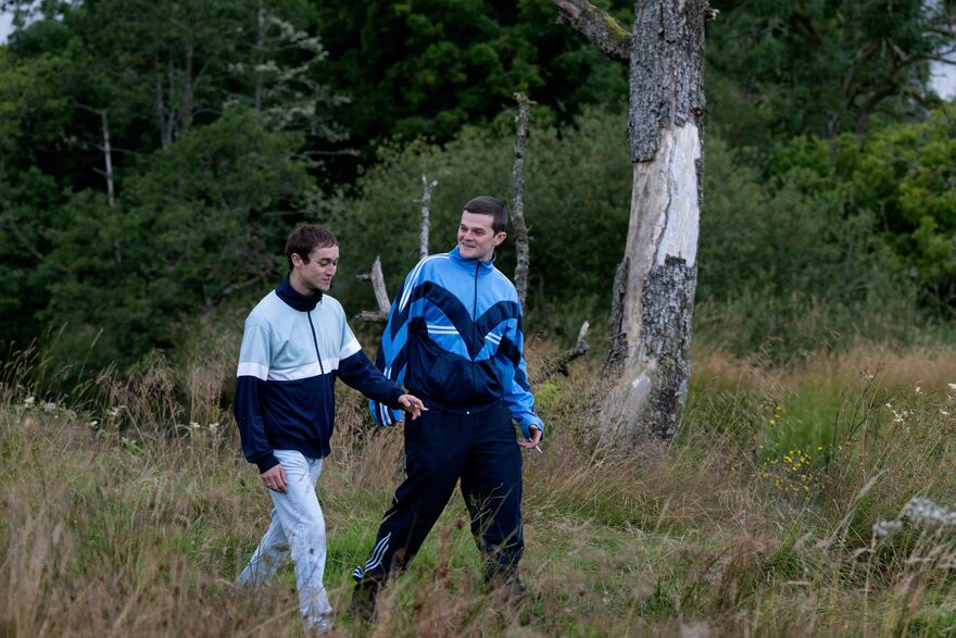 Two young men wearing tracksuits walk through a forest together laughing