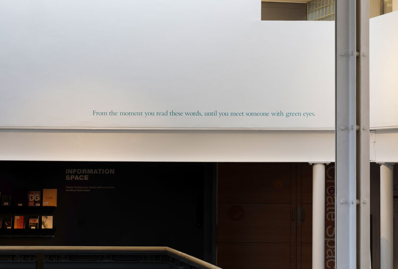 This image shows a text artwork in vinyl on the white wall external to the gallery, above head height. It reads