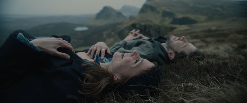 A man and woman lie on the grass on a hill together looking up at the sky and laughing.